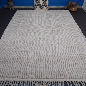 May include: Cream-colored rectangular rug with a textured, striped pattern and fringe along the bottom edge. The rug has a neutral color palette, suitable for various interior design styles. The rug is made of a soft material.