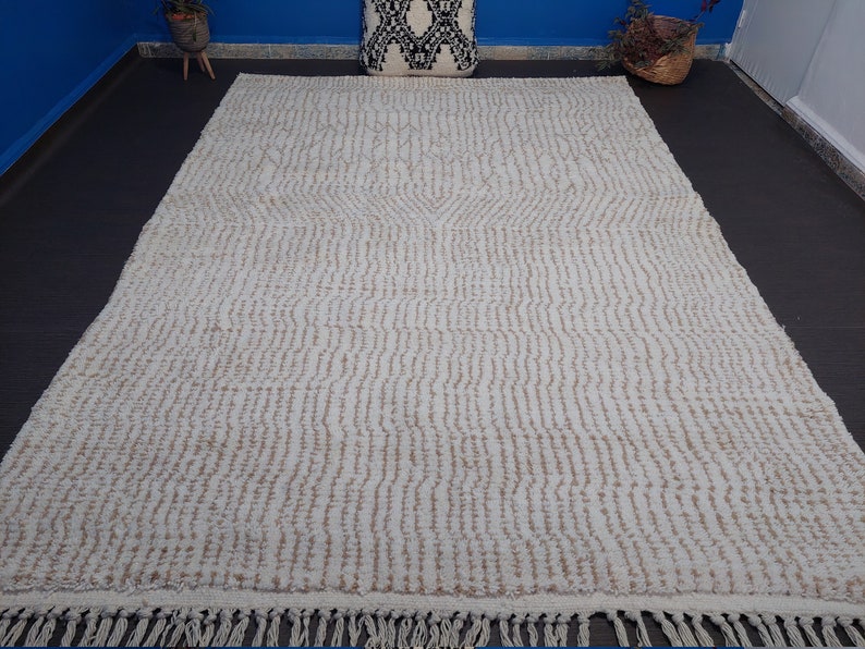 May include: Cream-colored rectangular rug with a textured, striped pattern and fringe along the bottom edge. The rug has a neutral color palette, suitable for various interior design styles. The rug is made of a soft material.