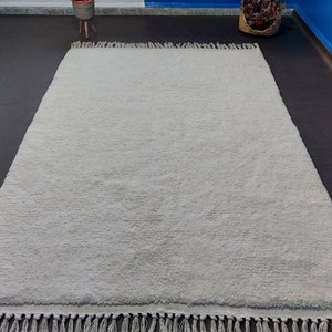 May include: A white shag rug with a fringe border. The rug is laid out on a dark grey floor.