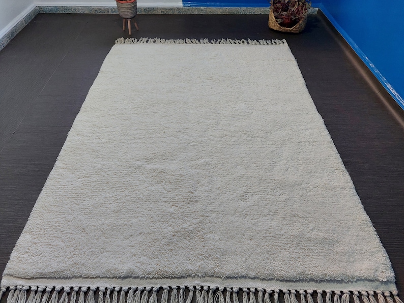 May include: A white shag rug with a fringe border. The rug is laid out on a dark grey floor.