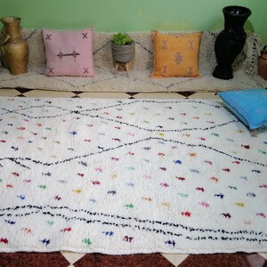 Puede incluir: Una alfombra marroqu&iacute; blanca con un patr&oacute;n de rombos negros y acentos de colores. La alfombra tiene flecos en los bordes y est&aacute; colocada sobre un suelo de baldosas.
