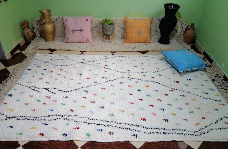 Puede incluir: Una alfombra marroqu&iacute; blanca con un patr&oacute;n de rombos negros y acentos de colores. La alfombra tiene flecos en los bordes y est&aacute; colocada sobre un suelo de baldosas.
