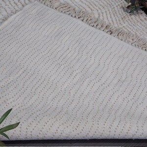 May include: Off-white woven rug with a textured, diagonal pattern. The rug has a fringed edge and a neutral color palette, suitable for various interior styles. The rug is folded over, revealing the underside.