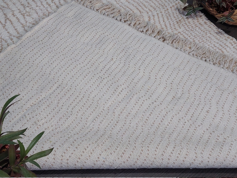 May include: Off-white woven rug with a textured, diagonal pattern. The rug has a fringed edge and a neutral color palette, suitable for various interior styles. The rug is folded over, revealing the underside.
