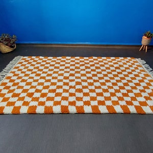 Op de afbeelding: Een rechthoekig tapijt met een dambordpatroon in oranje en wit. Het tapijt heeft franjes aan de korte uiteinden. Het ligt op een donkergrijze ondergrond, met een blauwe muur op de achtergrond.