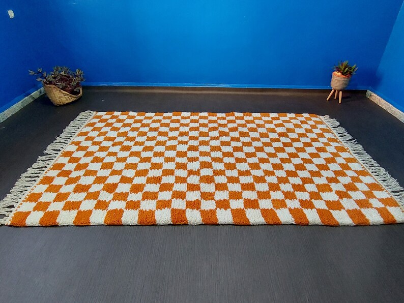 Op de afbeelding: Een rechthoekig tapijt met een dambordpatroon in oranje en wit. Het tapijt heeft franjes aan de korte uiteinden. Het ligt op een donkergrijze ondergrond, met een blauwe muur op de achtergrond.