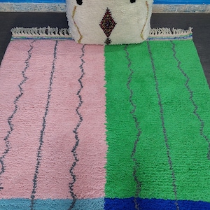 May include: A pink and green Moroccan rug with a blue and white border. The rug has a wavy pattern and is made of wool.