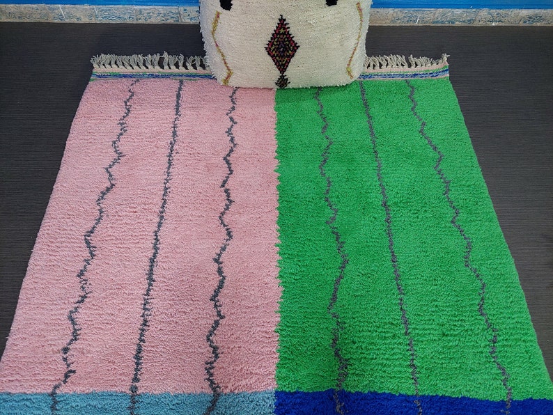 May include: A pink and green Moroccan rug with a blue and white border. The rug has a wavy pattern and is made of wool.