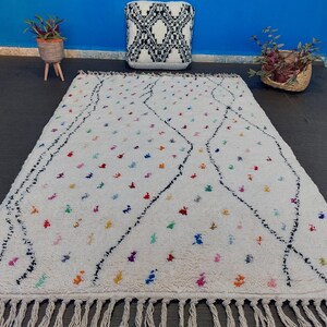 Puede incluir: Una alfombra marroqu&iacute; blanca con un patr&oacute;n de diamantes negros y acentos coloridos. La alfombra tiene un borde de flecos.