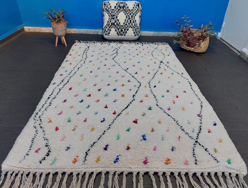 Puede incluir: Una alfombra marroqu&iacute; blanca con un patr&oacute;n de diamantes negros y acentos coloridos. La alfombra tiene un borde de flecos.