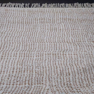 May include: Cream-colored rug with a textured, striped pattern. The rug has a fringe along one edge. The design features vertical lines of a light brown color against a cream background. This rug is suitable for various home decor styles.