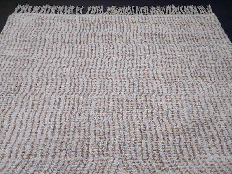 May include: Cream-colored rug with a textured, striped pattern. The rug has a fringe along one edge. The design features vertical lines of a light brown color against a cream background. This rug is suitable for various home decor styles.