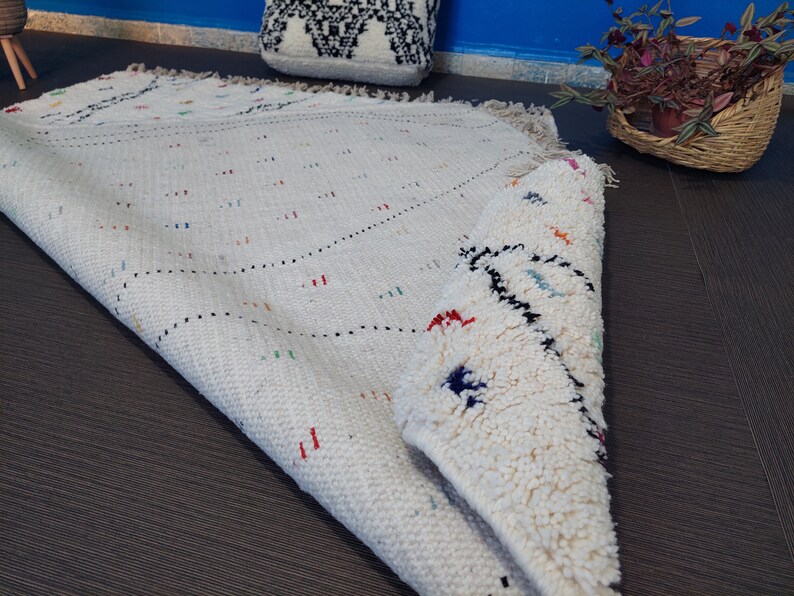 Puede incluir: Una alfombra marroqu&iacute; blanca con un patr&oacute;n geom&eacute;trico en hilos negros y coloridos. La alfombra est&aacute; doblada por la mitad y tiene una textura esponjosa.