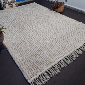 May include: Cream-colored rectangular rug with a textured, striped pattern. The rug has a fringe along one edge. The rug is placed on a dark surface, with plants and decorative items in the background.