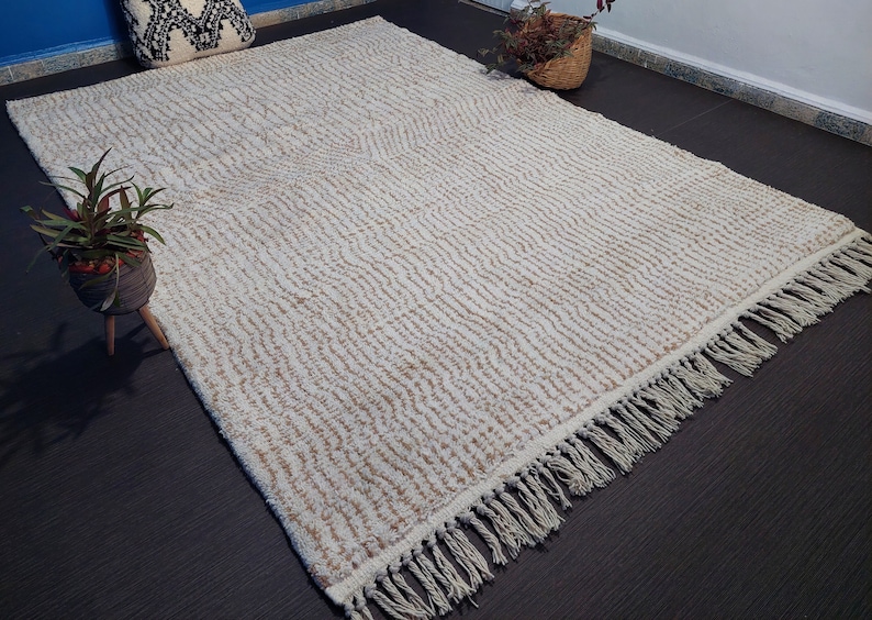 May include: Cream-colored rectangular rug with a textured, striped pattern. The rug has a fringe along one edge. The rug is placed on a dark surface, with plants and decorative items in the background.