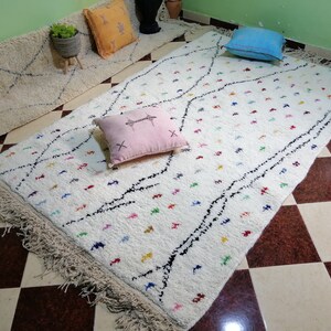 Puede incluir: Alfombra marroqu&iacute; blanca con acentos negros y coloridos. La alfombra tiene un borde de flecos y est&aacute; colocada sobre un suelo a cuadros.