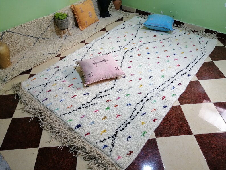 Puede incluir: Alfombra marroqu&iacute; blanca con acentos negros y coloridos. La alfombra tiene un borde de flecos y est&aacute; colocada sobre un suelo a cuadros.