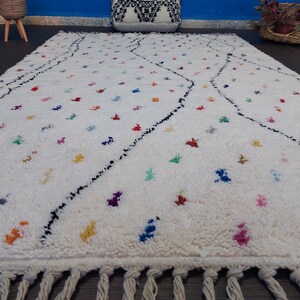 Puede incluir: Una alfombra blanca con un patr&oacute;n geom&eacute;trico blanco y negro y acentos de colores. La alfombra tiene un borde de flecos.