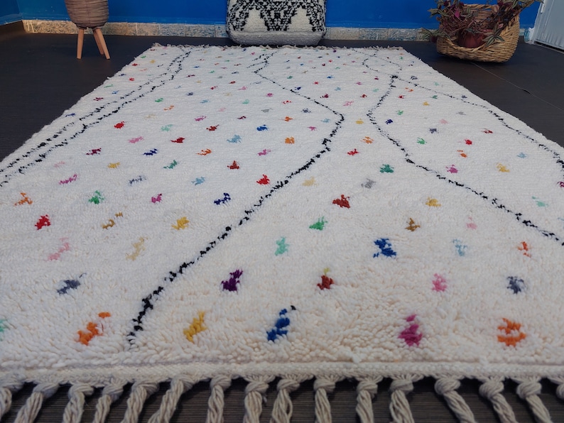 Puede incluir: Una alfombra blanca con un patr&oacute;n geom&eacute;trico blanco y negro y acentos de colores. La alfombra tiene un borde de flecos.