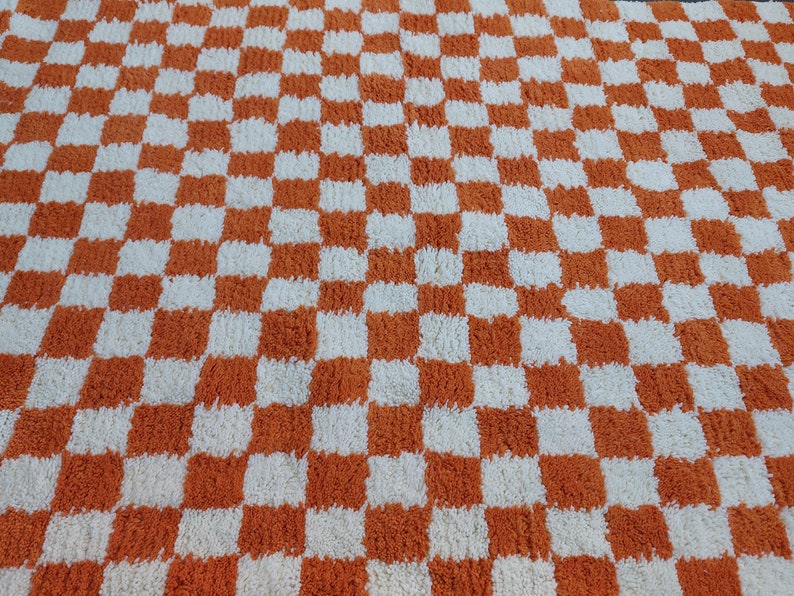 Op de afbeelding: Een geruit tapijt met een herhalend patroon van oranje en witte vierkanten. Het tapijt heeft een zacht, getextureerd uiterlijk, wat wijst op een zacht materiaal. Het ontwerp is een klassiek dambordpatroon.