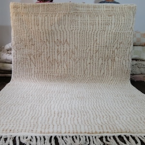 May include: A large, off-white rug with a textured, vertical stripe pattern and a fringe along the bottom edge. The rug has a geometric design in a lighter brown color. The rug is made of a thick, woven material.