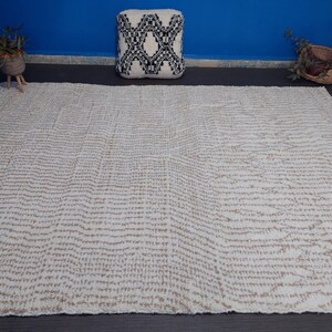 May include: A large, rectangular rug with a cream-colored background and a repeating pattern of thin, horizontal, tan lines. The rug has a fringed edge and is placed on a dark surface. A decorative pillow and plants are in the background.