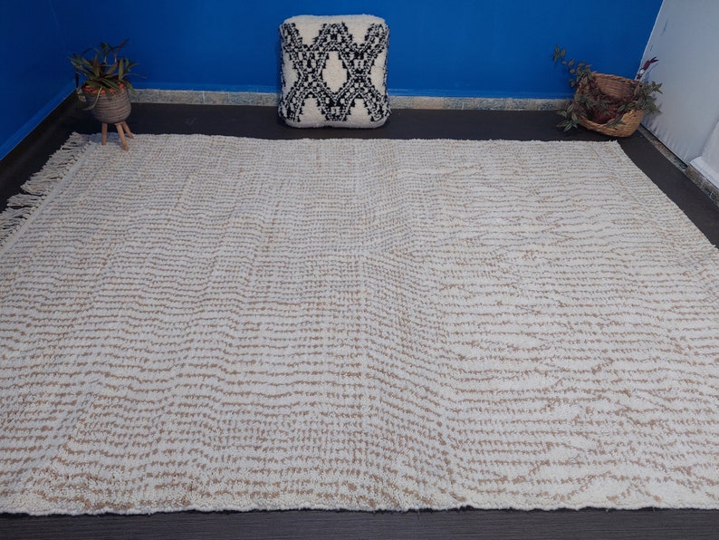 May include: A large, rectangular rug with a cream-colored background and a repeating pattern of thin, horizontal, tan lines. The rug has a fringed edge and is placed on a dark surface. A decorative pillow and plants are in the background.