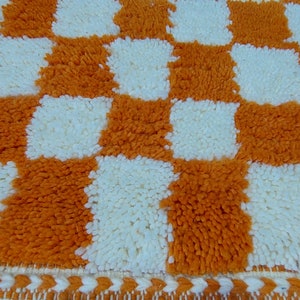 Op de afbeelding: Close-up van een getextureerd, oranje en wit geblokt tapijt. Het tapijt heeft een herhalend patroon van oranje en witte vierkanten. De randen van het tapijt hebben een decoratieve rand met een zigzagpatroon.