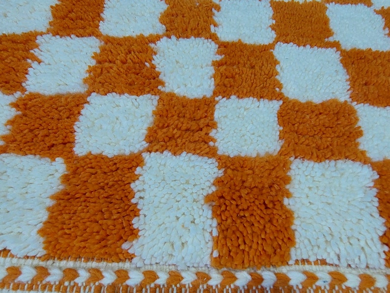 Op de afbeelding: Close-up van een getextureerd, oranje en wit geblokt tapijt. Het tapijt heeft een herhalend patroon van oranje en witte vierkanten. De randen van het tapijt hebben een decoratieve rand met een zigzagpatroon.