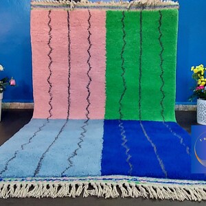 May include: A Moroccan rug with a geometric pattern in pink, green, blue, and light blue. The rug has a fringe border.