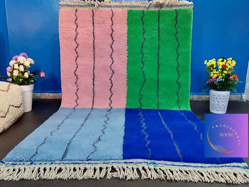 May include: A Moroccan rug with a geometric pattern in pink, green, blue, and light blue. The rug has a fringe border.