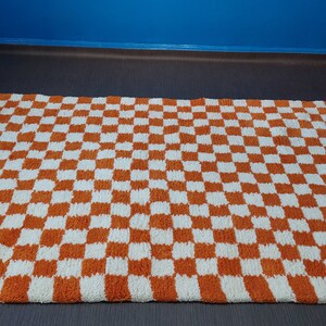 Op de afbeelding: Een rechthoekig tapijt met een dambordpatroon in oranje en wit. Het tapijt heeft een gefranjerde rand en ligt op een donkere ondergrond. Het ontwerp van het tapijt is een herhalend patroon van vierkanten.