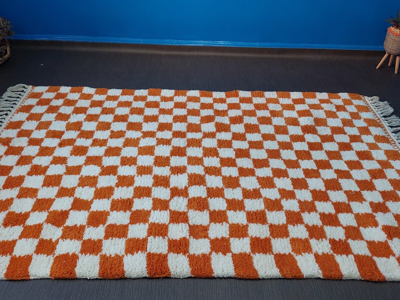 Op de afbeelding: Een rechthoekig tapijt met een dambordpatroon in oranje en wit. Het tapijt heeft een gefranjerde rand en ligt op een donkere ondergrond. Het ontwerp van het tapijt is een herhalend patroon van vierkanten.