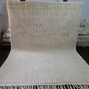 May include: Cream-colored rug with a textured, vertical striped pattern and a fringe along the bottom edge. The rug has a neutral color palette, with light brown accents. The rug is made of a soft material.