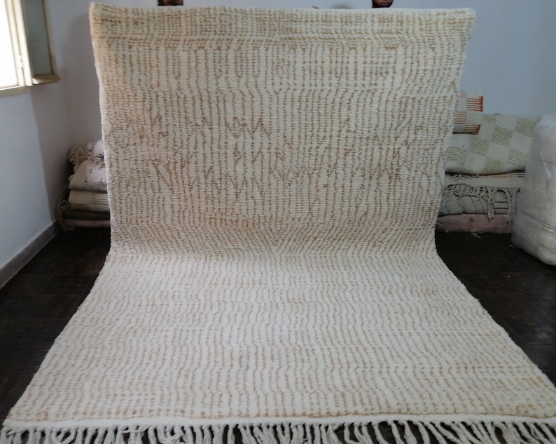 May include: Cream-colored rug with a textured, vertical striped pattern and a fringe along the bottom edge. The rug has a neutral color palette, with light brown accents. The rug is made of a soft material.