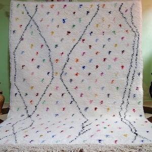 Puede incluir: Una alfombra marroqu&iacute; blanca con un patr&oacute;n de diamantes y acentos coloridos. La alfombra tiene un borde de flecos y est&aacute; hecha de lana.
