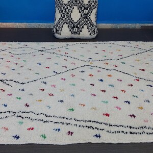 Puede incluir: Una alfombra marroqu&iacute; blanca con un patr&oacute;n de diamantes negros y acentos coloridos. La alfombra tiene un borde de flecos y est&aacute; hecha de lana.