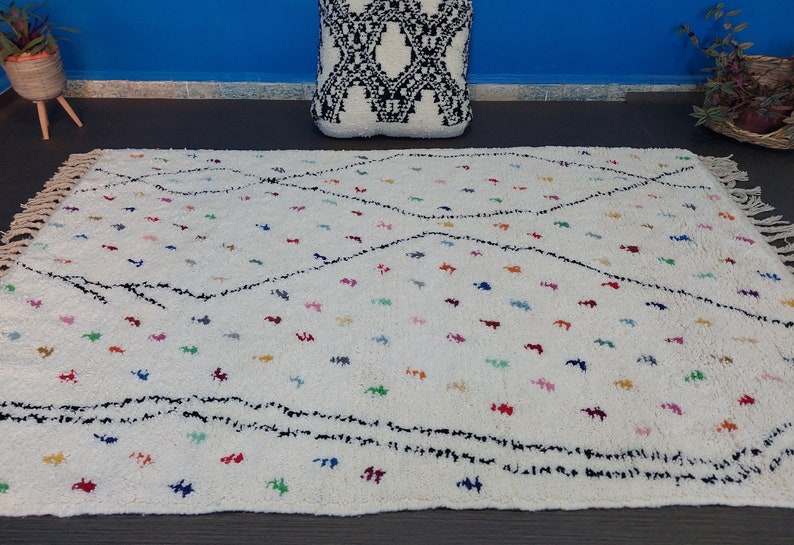 Puede incluir: Una alfombra marroqu&iacute; blanca con un patr&oacute;n de diamantes negros y acentos coloridos. La alfombra tiene un borde de flecos y est&aacute; hecha de lana.
