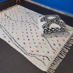 Puede incluir: Una alfombra marroqu&iacute; blanca con patrones geom&eacute;tricos negros y coloridos y flecos. Una almohada con estampado en blanco y negro est&aacute; encima de la alfombra.