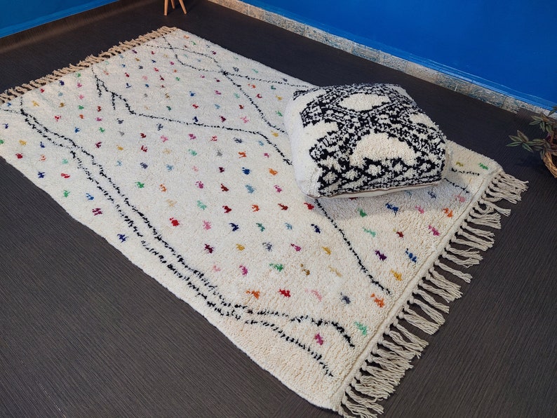 Puede incluir: Una alfombra marroqu&iacute; blanca con patrones geom&eacute;tricos negros y coloridos y flecos. Una almohada con estampado en blanco y negro est&aacute; encima de la alfombra.