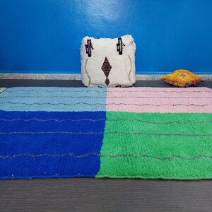May include: A colorful Moroccan rug with four squares in blue, green, pink, and light blue. The rug is on a dark wood floor with a white pillow with a geometric pattern and a yellow pillow on top of it.