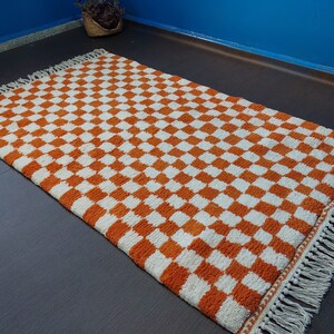 Op de afbeelding: Rechthoekig vloerkleed met een oranje en wit dambordpatroon. Het kleed heeft een cr&egrave;mekleurige franje langs de randen. Het patroon herhaalt zich. Het kleed lijkt gemaakt van een geweven materiaal en is geschikt voor diverse ruimtes.