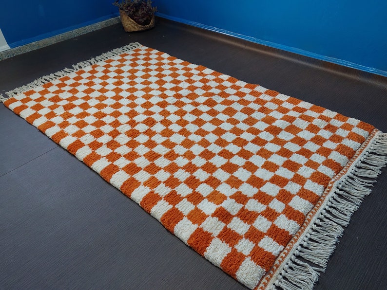 Op de afbeelding: Rechthoekig vloerkleed met een oranje en wit dambordpatroon. Het kleed heeft een cr&egrave;mekleurige franje langs de randen. Het patroon herhaalt zich. Het kleed lijkt gemaakt van een geweven materiaal en is geschikt voor diverse ruimtes.