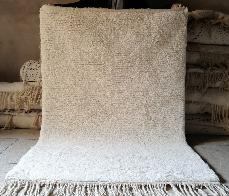 May include: A white, fluffy rug with a fringe border. The rug is made of a soft, textured material and has a subtle sheen.