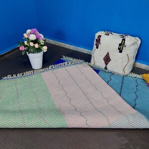 May include: A multi-colored woven rug with green, pink, and blue sections. The rug has a fringe border and is laid out on a dark floor. A white pouf with a geometric pattern sits on the rug.