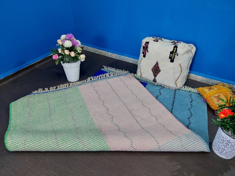 May include: A multi-colored woven rug with green, pink, and blue sections. The rug has a fringe border and is laid out on a dark floor. A white pouf with a geometric pattern sits on the rug.