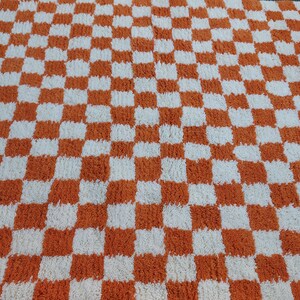 Op de afbeelding: Een geruit tapijt met een herhalend patroon van oranje en witte vierkanten. Het tapijt heeft een zacht, getextureerd uiterlijk, wat wijst op een zacht materiaal. Het ontwerp is een klassiek dambordpatroon.