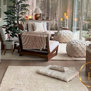 May include: A cozy living room with a white rug, a wooden chair with a pink and white zebra print throw blanket, two white poufs, a wicker basket, and a potted plant. The room has large windows and a light green wall.