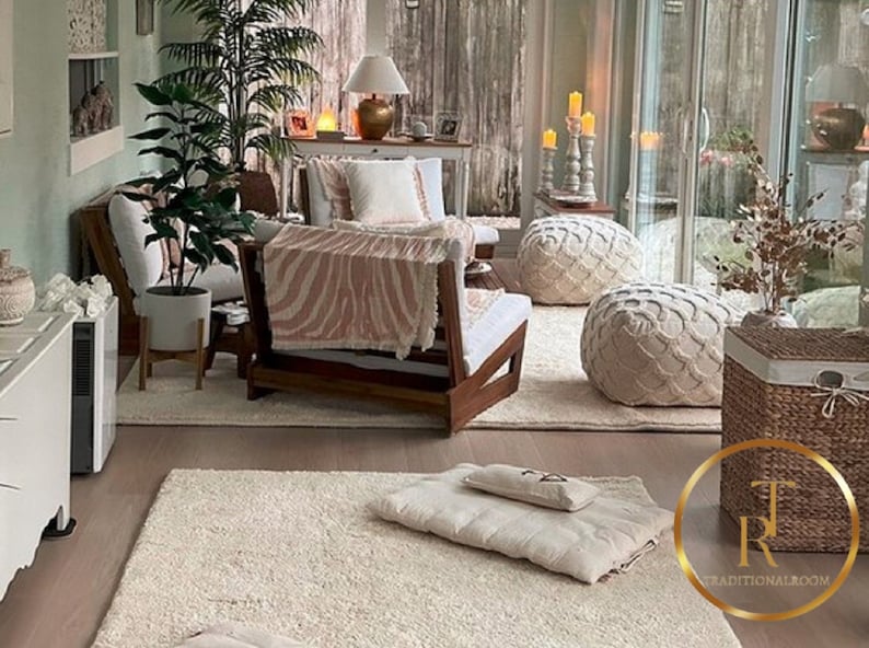 May include: A cozy living room with a white rug, a wooden chair with a pink and white zebra print throw blanket, two white poufs, a wicker basket, and a potted plant. The room has large windows and a light green wall.