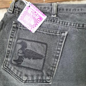 May include: A pair of charcoal gray denim jeans featuring a black square patch on the back pocket with a dark silhouette of a loon bird. A bright pink, square-shaped paper tag with a white geometric pattern is attached to the waistband.